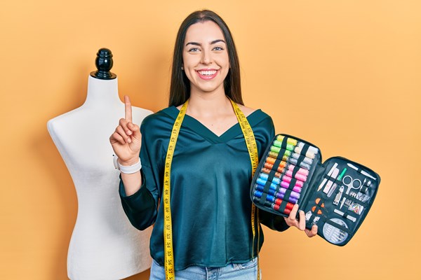 alt="jonge dame houdt koffertje vast met naaigereedschap en heeft lintmeter rond haar nek. Ze glimlacht en achter haar staat een paspop"